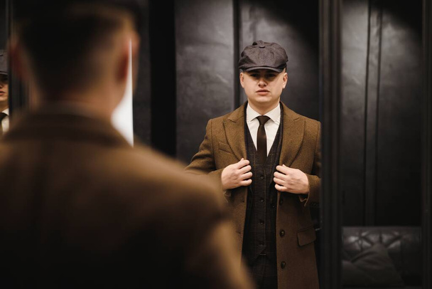 A man posing in the image of an English retro gangster of the 1920s dressed in a coat, suit and flat cap in Peaky blinders style. - Photo, image