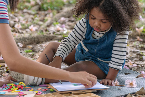 小さな巻き毛の混合レースの女の子は葉と芝生の上に座って、公園内の手の母親と何かを学ぶ。親が庭で教える間、幸せな子供たちの夏の活動. - 写真・画像