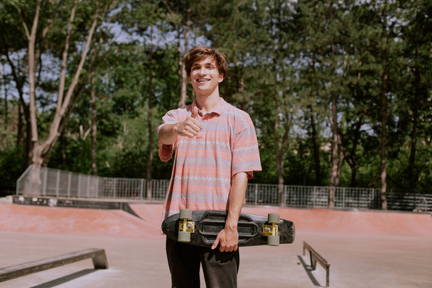 Primer plano de la cámara increíble hombre gran sonrisa hermosa en un moderno parque de skate sosteniendo su monopatín y sintiéndose feliz de que tiene traje hipster - Foto, Imagen