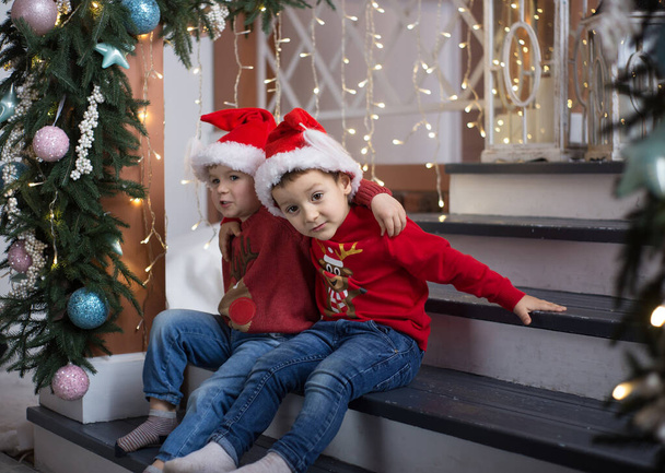 dos chicos sonrientes alegres lindos de cuatro años en sombreros de Santa y suéteres rojos se sientan en las escaleras y abrazan, contra el fondo de las luces de Navidad de guirnaldas - Foto, imagen