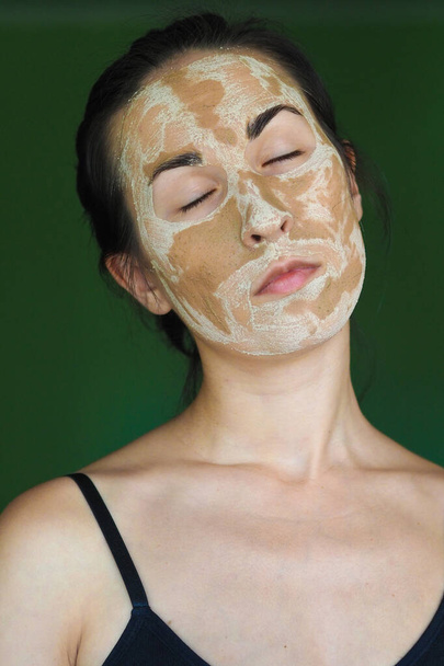 junge brünette Frau mit einer hausgemachten Maske aus Ton auf dem Gesicht mit geschlossenen Augen - Foto, Bild