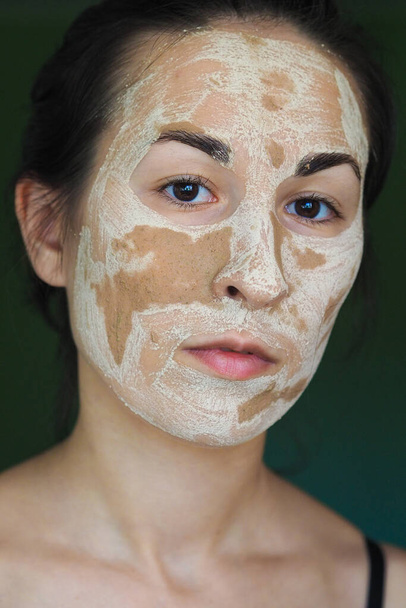  νεαρή μελαχρινή γυναίκα με μια πήλινη σπιτική μάσκα στο πρόσωπό της κοιτάζει μέσα στο πλαίσιο - Φωτογραφία, εικόνα
