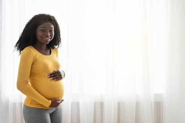 Beautiful pregnant african woman hugging her big belly at home - Photo, Image