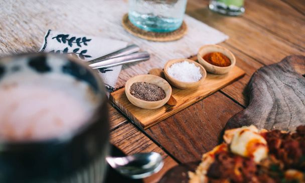Z góry miski z solą czarny pieprz i czerwony pieprz umieszczone na drewnianym stole w kawiarni w pobliżu filiżanki kawy - Zdjęcie, obraz