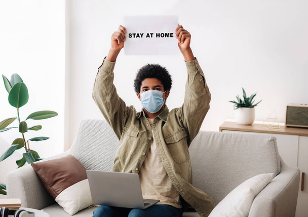 African American teenager in medical mask lifting STAY AT HOME sign above his head, indoors - Photo, Image