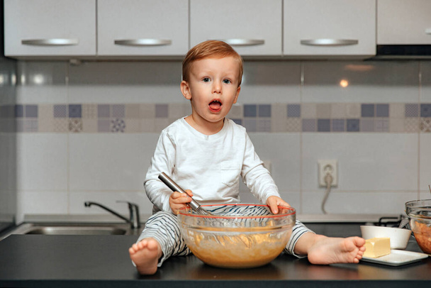 Cute blond boy using whisker while mixing flour and eggs in bowl in kitchen environment. - Foto, imagen