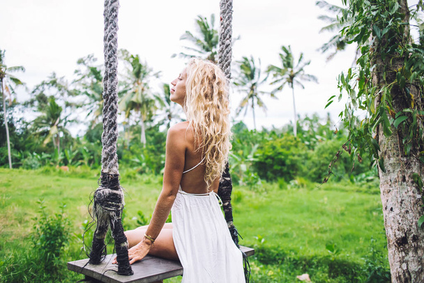 Side view of blonde modern curly young woman in white summer outfit sitting on swing in grassy palm garden having holiday looking away  - Fotó, kép