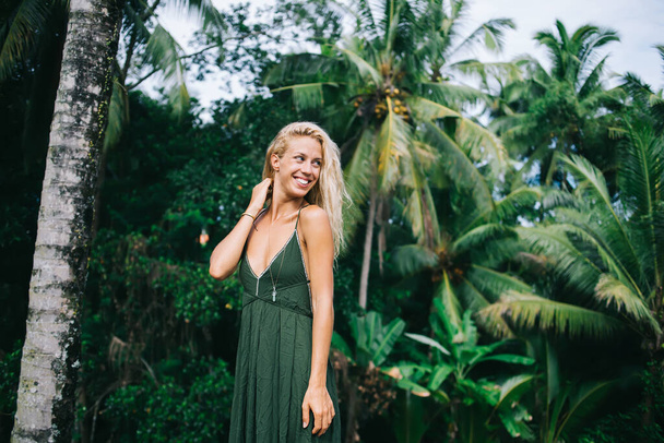 Beautiful blonde woman in green trendy dress with neckline standing in forest by palm trees smiling and looking over shoulder - Valokuva, kuva