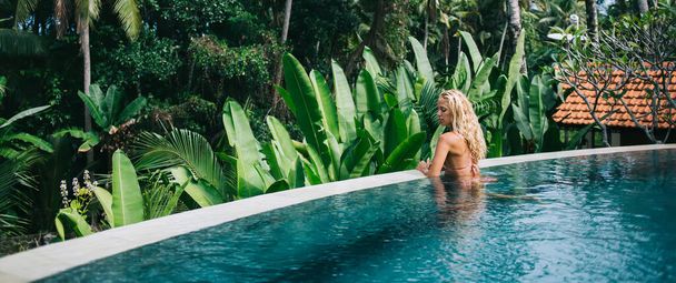 Side view of pensive blonde woman chilling in luxury pool by tropical plants and palm trees and looking down on daytime - Fotó, kép