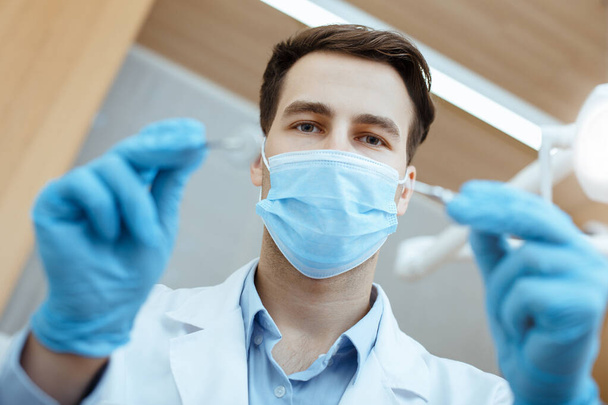 Junge attraktive Zahnärztin in weißem Kittel, Schutzmaske und Gummihandschuhen behandelt Patienten mit Zähnen - Foto, Bild