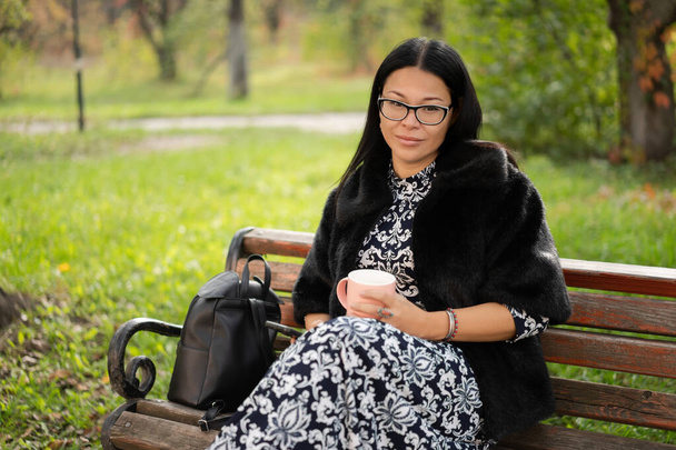 Smiling woman holds coffee cup relaxing outdoors on sunny day. Beautiful Asian lady wearing fur coat with cup of hot drink resting on bench in city garden - 写真・画像