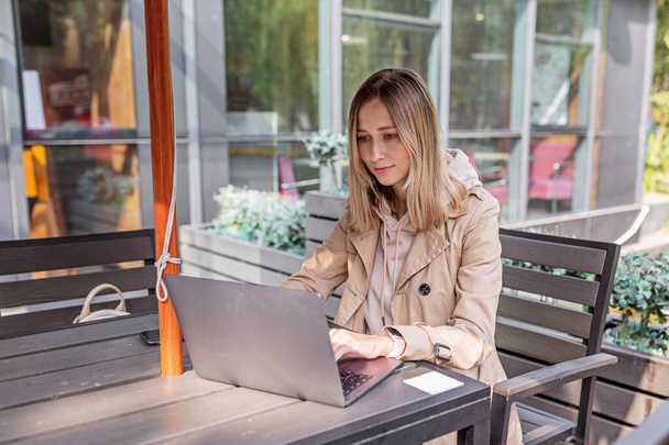 Young Caucasian business woman with blonde hair working on laptop in outdoor cafe. College student using technology , online education, freelance . High quality photo - Фото, изображение