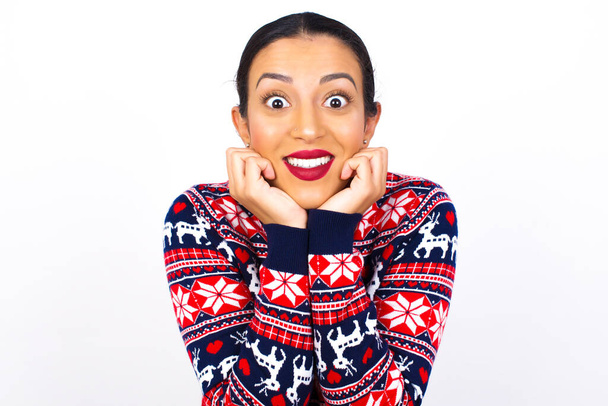 Marzycielska młoda piękna kobieta ubrana w świąteczny sweter pod białą ścianą, trzyma ręce przyciśnięte do podbródka, wygląda szczęśliwie, ma zębaty uśmiech.  - Zdjęcie, obraz
