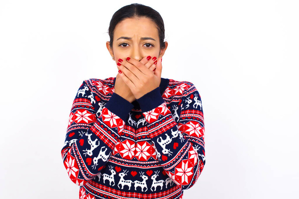 Zdenerwowana młoda piękna kobieta nosząca świąteczny sweter pod białą ścianą, zakrywająca usta obiema dłońmi, aby zapobiec krzykom, po zobaczeniu lub usłyszeniu czegoś złego.  - Zdjęcie, obraz