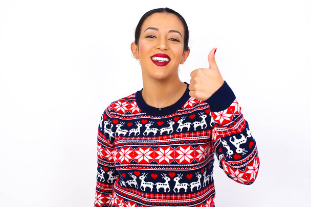 Young beautiful  woman wearing Christmas sweater against white wall, giving thumb up gesture, good Job! Positive human emotion facial expression body language. - Photo, Image