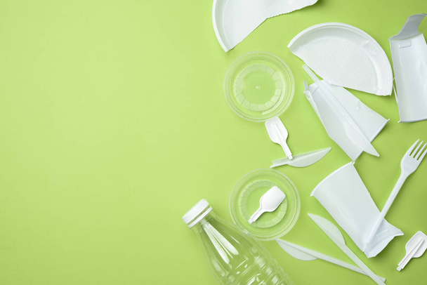 utensílios de mesa de plástico branco quebrado, garrafa transparente no fundo verde, conceito de poluição e meio ambiente, vista superior - Foto, Imagem