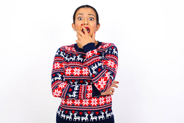 Joven mujer árabe hermosa usando suéter de Navidad contra la pared blanca cubriendo la boca con las manos asustadas de algo o alguien mordiendo clavos - Foto, imagen