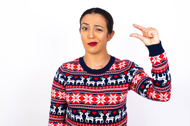 Perturbado Jovem bela mulher árabe vestindo camisola de Natal contra formas de parede branca pouco gesto com a mão demonstra algo muito pequeno tamanho. Não muito. - Foto, Imagem