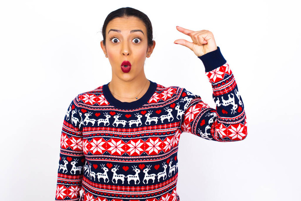 Шокированная молодая красивая арабская женщина в рождественском свитере на белой стене показывает что-то мало с руками, демонстрирует размер, открывает рот от удивления. Концепция измерения. - Фото, изображение