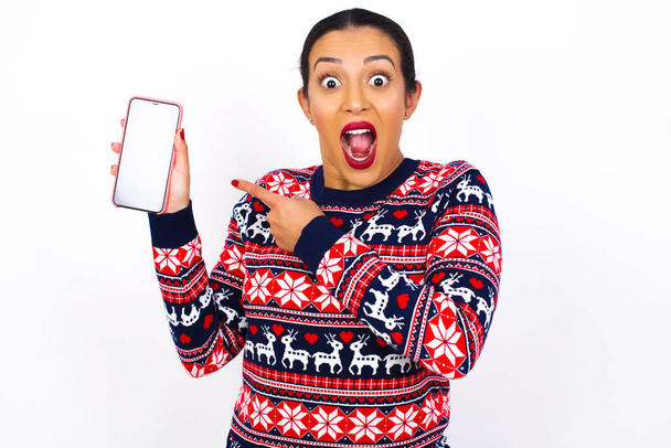 Emocionado Joven mujer árabe hermosa con suéter de Navidad sosteniendo y señalando con el dedo en el teléfono inteligente con pantalla en blanco. Concepto de publicidad. - Foto, imagen