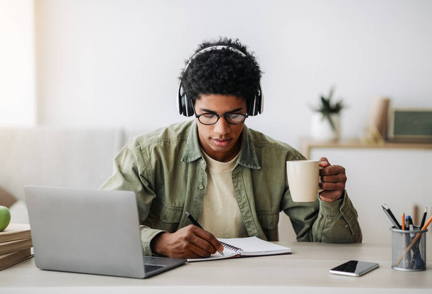 Zajęty nastolatek pijący kawę podczas lekcji online na laptopie, zapisujący informacje w notatniku, w pomieszczeniach - Zdjęcie, obraz