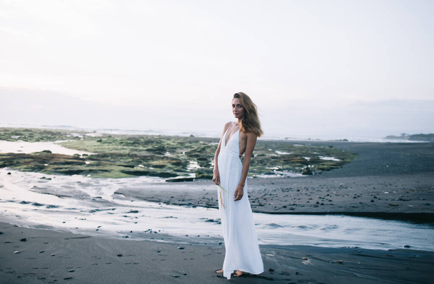 砂浜の海岸に立つ白いドレスを着た若い裸足の女性の完全な長さとバリの夏休みを楽しみながらカメラを見て - 写真・画像