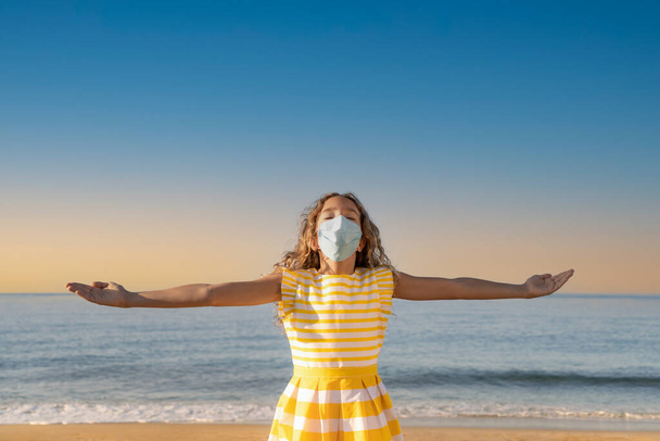 Happy child wearing medical mask outdoor against blue sky background. Girl enjoying by sea at summer. Coronavirus pandemic and healthy lifestyle concept - Foto, imagen