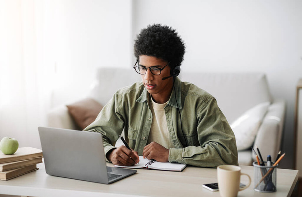 Adolescente negro industrial en auriculares estudiando remotamente desde casa, escribiendo en un cuaderno - Foto, imagen