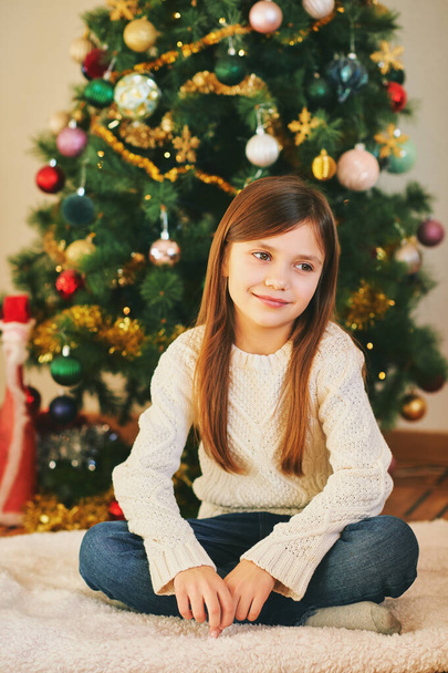 Porträt eines süßen kleinen Mädchens, das neben dem Weihnachtsbaum sitzt und weißen Pullover trägt, Winterurlaub - Foto, Bild