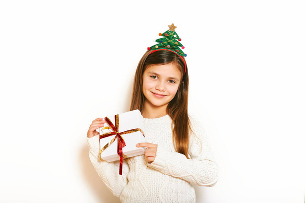 Studio Christmas portrait of young sweet little girl on white background, wearing winter pullover and xmas tree haeadband, holding packed present - Zdjęcie, obraz