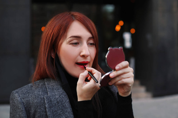 Roodharige blanke zakenvrouw die naar een kleine zakspiegel kijkt en rode lippenstift aanbrengt na het werk in de buurt van haar kantoor voor het daten. Snelle make-over voor de vergadering. Make-up concept. - Foto, afbeelding