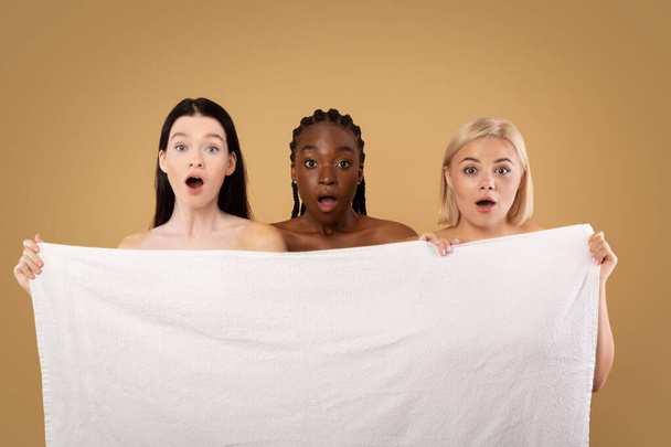 Τρεις συναισθηματικές πολυ-εθνοτικές γυναίκες που κρύβονται πίσω από την πετσέτα μπάνιου με σοκαρισμένη έκφραση προσώπου - Φωτογραφία, εικόνα