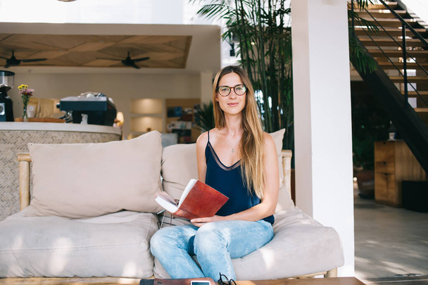 Focused female entrepreneur in casual clothes and eyeglasses reading notes in planner while resting on comfortable couch in modern cafe - Foto, Imagem