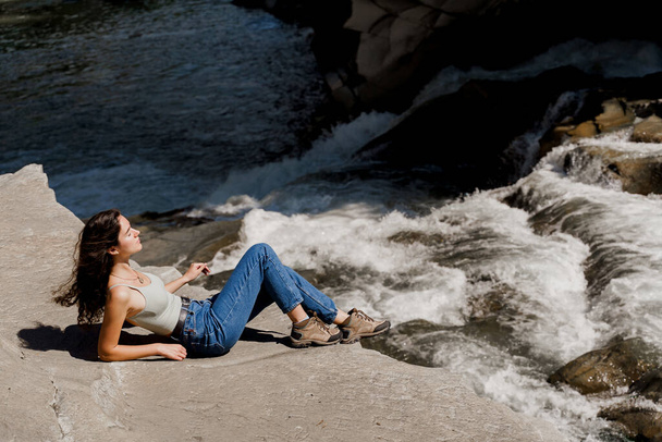 Traveler girl is sitting on the rock near waterfall and looking toward. Travelling in Karpathian mountains. Cascade waterfall. Beautiful landscape - Photo, Image