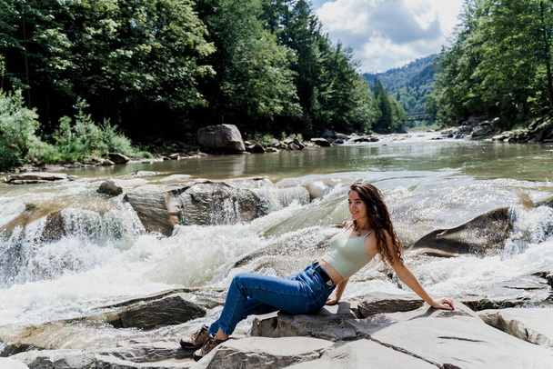 Мандрівниця сидить на скелі біля водоспаду і дивиться в бік. Подорожував Карпатськими горами. Каскадний водоспад. Чудовий краєвид - Фото, зображення