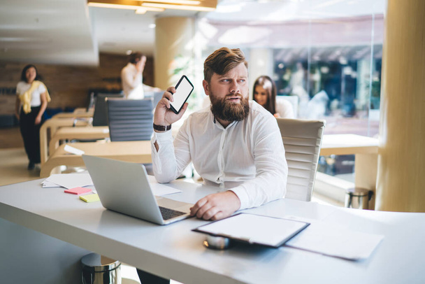 Serious male entrepreneur in elegant wear sitting at table with laptop and using mobile phone while thinking of startup ideas and looking away - 写真・画像