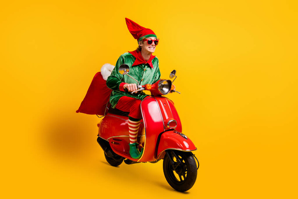 Portrait of his he nice attractive cheerful cheery funny guy elf riding moped delivering sack gifts December Eve Noel sale isolated over bright vivid shine vibrant yellow color background - Zdjęcie, obraz