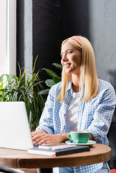 blonde freelancer smiling while typing on laptop in cafe on blurred foreground - Photo, Image