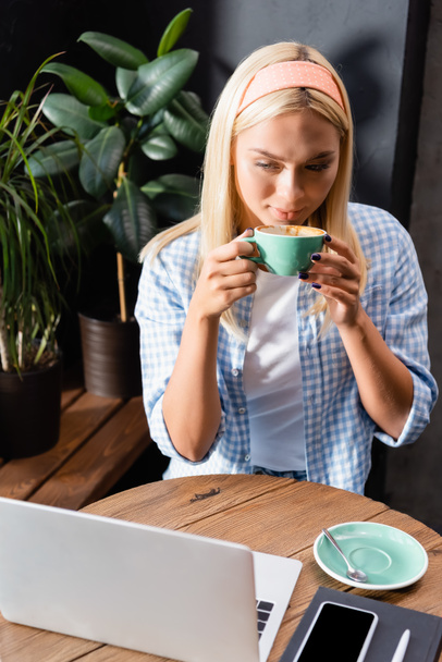 blonde freelancer drinking coffee near laptop and smartphone with blank screen in cafe - Photo, Image