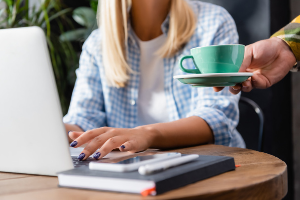 bijgesneden weergave van ober holding kopje koffie in de buurt van freelancer typen op laptop in cafe - Foto, afbeelding