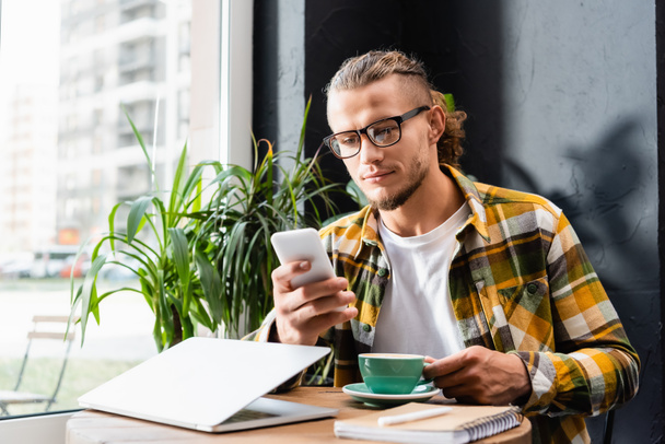 stylish freelancer in plaid shirt and eyeglasses chatting on smartphone near cup of coffee and laptop, blurred foreground - Foto, Imagem