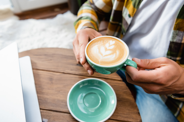 обрізаний вид людини, що сидить у кафе і тримає чашку кави з латте мистецтвом
 - Фото, зображення