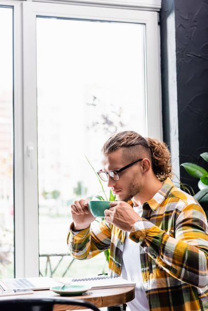 elegante freelancer en gafas y camisa a cuadros sosteniendo taza de café mientras está sentado en la cafetería cerca de la computadora portátil - Foto, Imagen