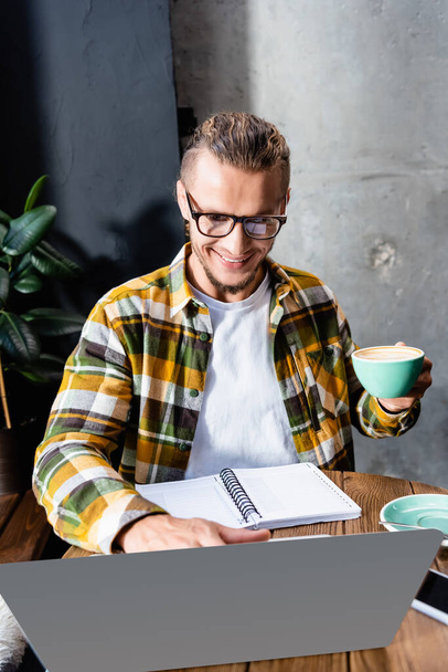 Ekose gömlekli ve gözlüklü neşeli serbest yazar kahve fincanı tutuyor ve kafede dizüstü bilgisayar kullanıyor. - Fotoğraf, Görsel