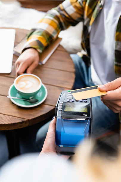 vista recortada del camarero con terminal de pago y el hombre con tarjeta de crédito sentado en la mesa con taza de café - Foto, Imagen