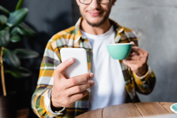partial view of freelancer messaging on smartphone while holding cup of coffee, blurred background - 写真・画像