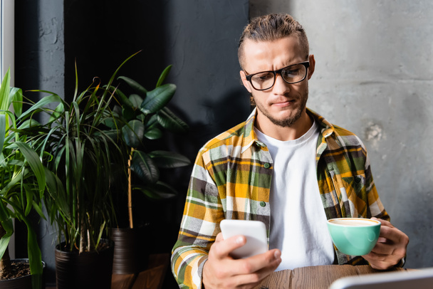 thoughtful man in plaid shirt and eyeglasses messaging on smartphone while holding cup of coffee in cafe - Foto, Bild