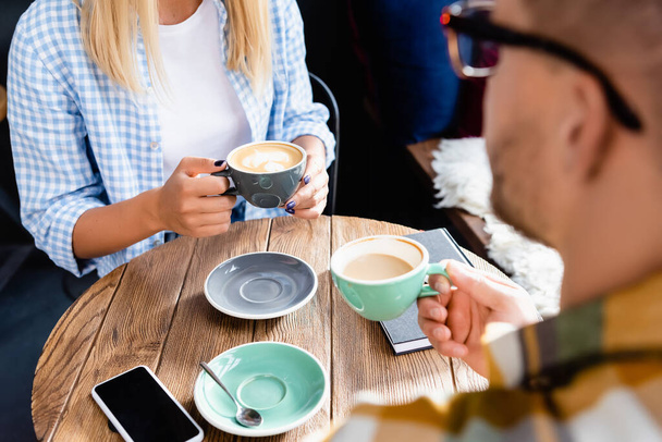 vista recortada de la mujer en camisa a cuadros y el hombre en primer plano borrosa beber café en la cafetería - Foto, Imagen