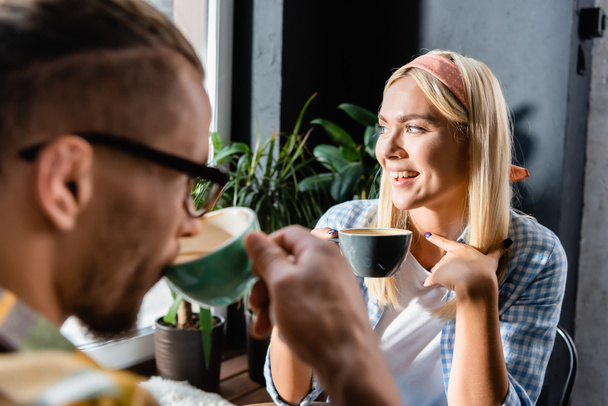 alegre joven mujer sosteniendo taza cerca amigo beber café en borrosa primer plano - Foto, Imagen