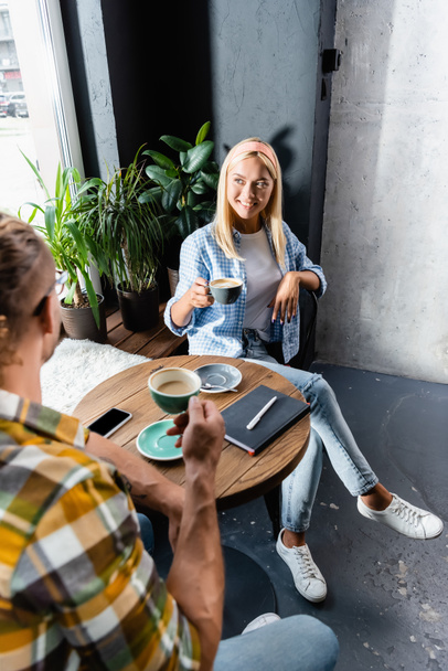 Lächelnde blonde Frau mit einer Tasse Kaffee, während sie mit einem Freund im Café sitzt - Foto, Bild